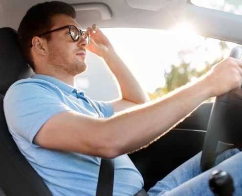 Man with glasses, wearing a blue shirt and jeans sitting in a car driving.